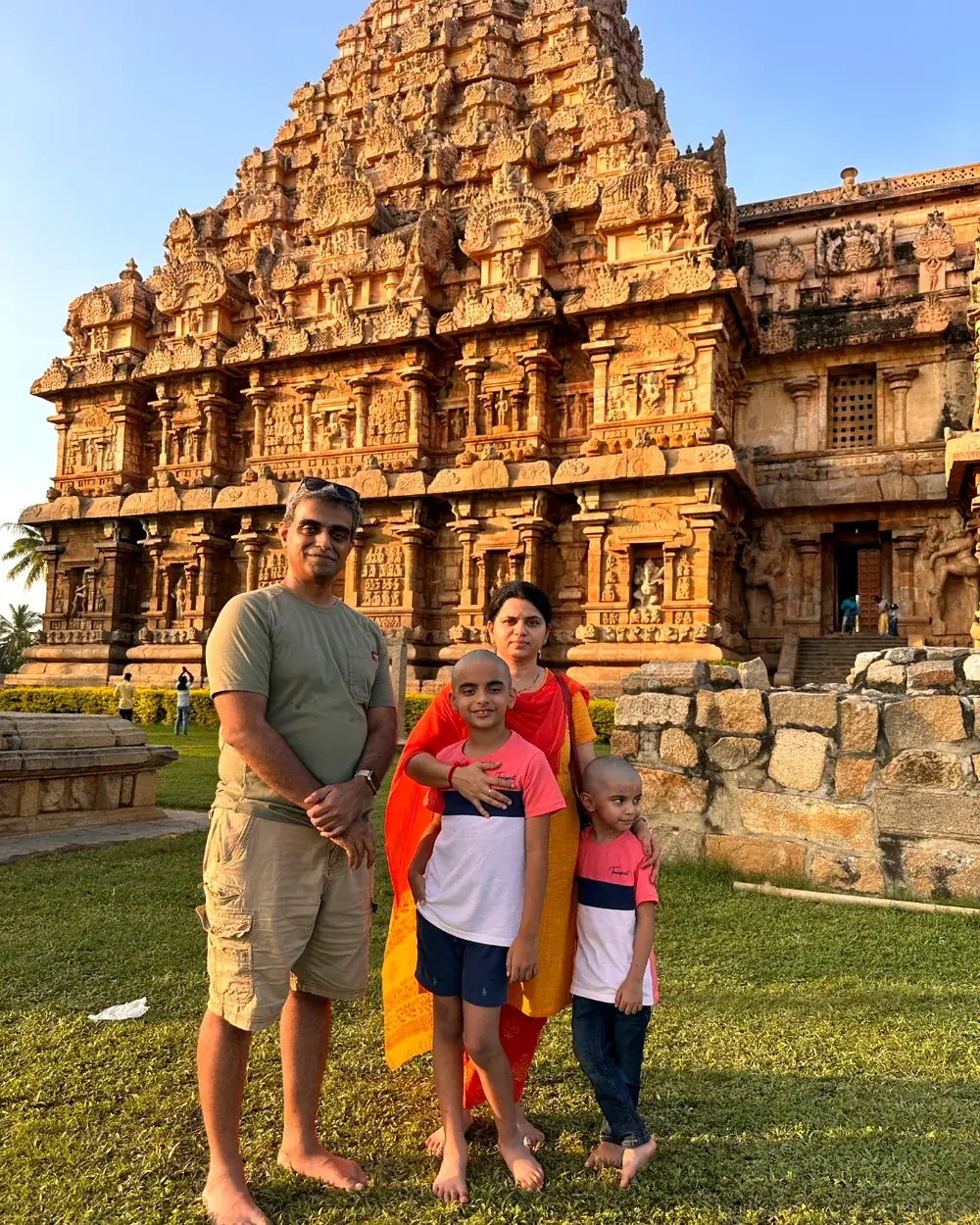 The family standing on the lawn in front of a great granite Chola temple vimana at golden hour