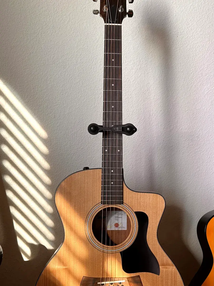 A Taylor acoustic-electric guitar on a stand against a white wall in slatted afternoon light
