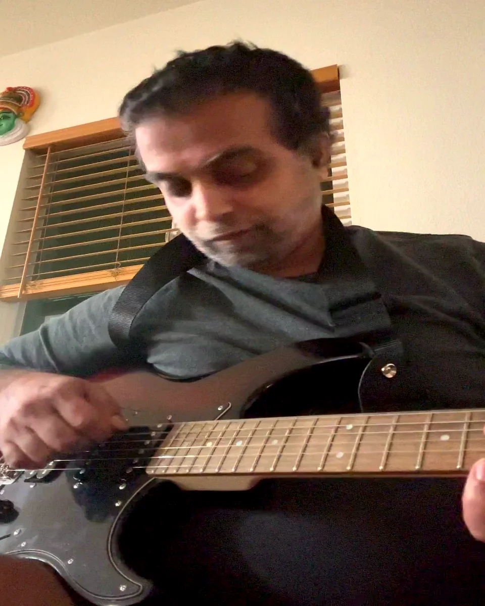 Bhargav Vivekanandan playing his black electric guitar at home, fretting hand on the neck
